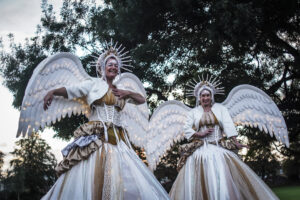 2 stilt walkers dressed as angels smile as they walk through a park