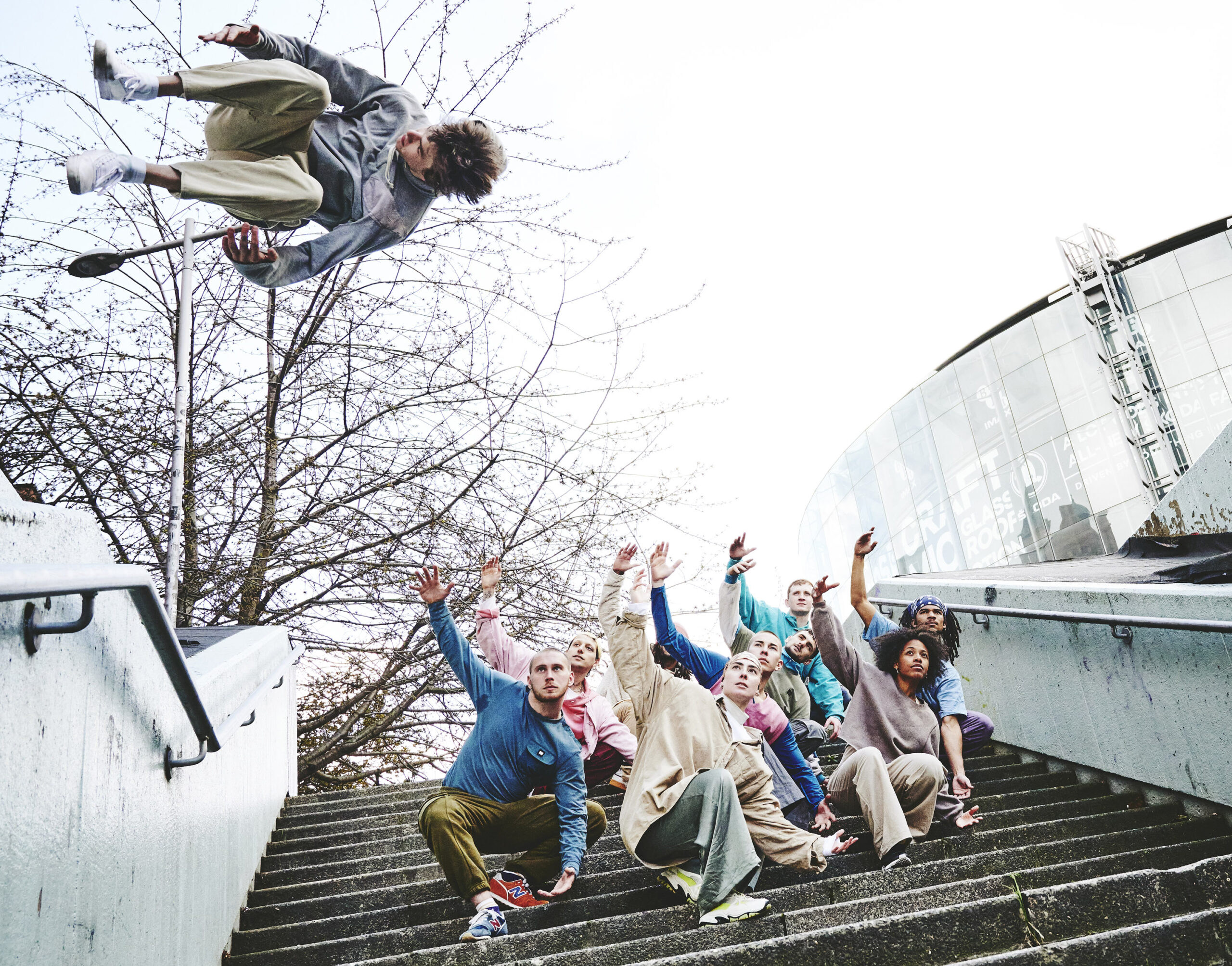 9 young people crouch on concrete steps under a grey sky. Their right arms are lifted up. A 10th person hovers sideways in mid-air with legs tucked in, halfway through a jump
