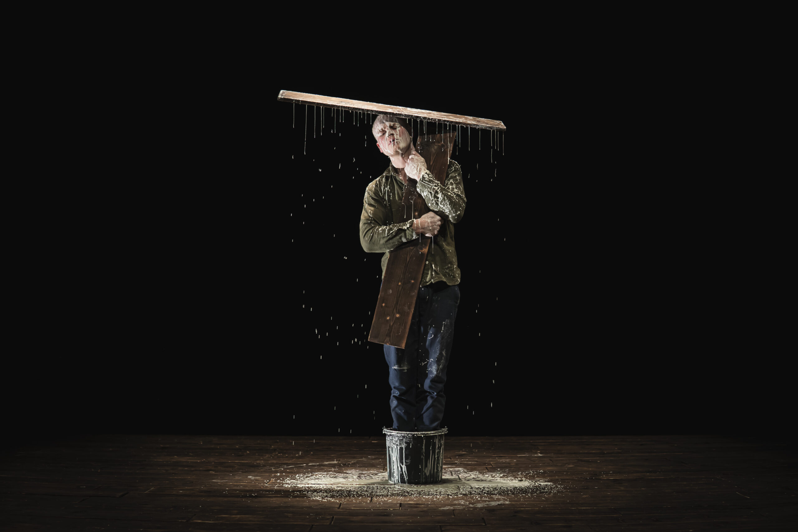 This is a photo of a spot-lit man standing in a black bucket holding up a plank of wood over his head which is covered in glue> The glue is dripping all over his head, face, clothes and the floor, creating a white pool on the stage around him.