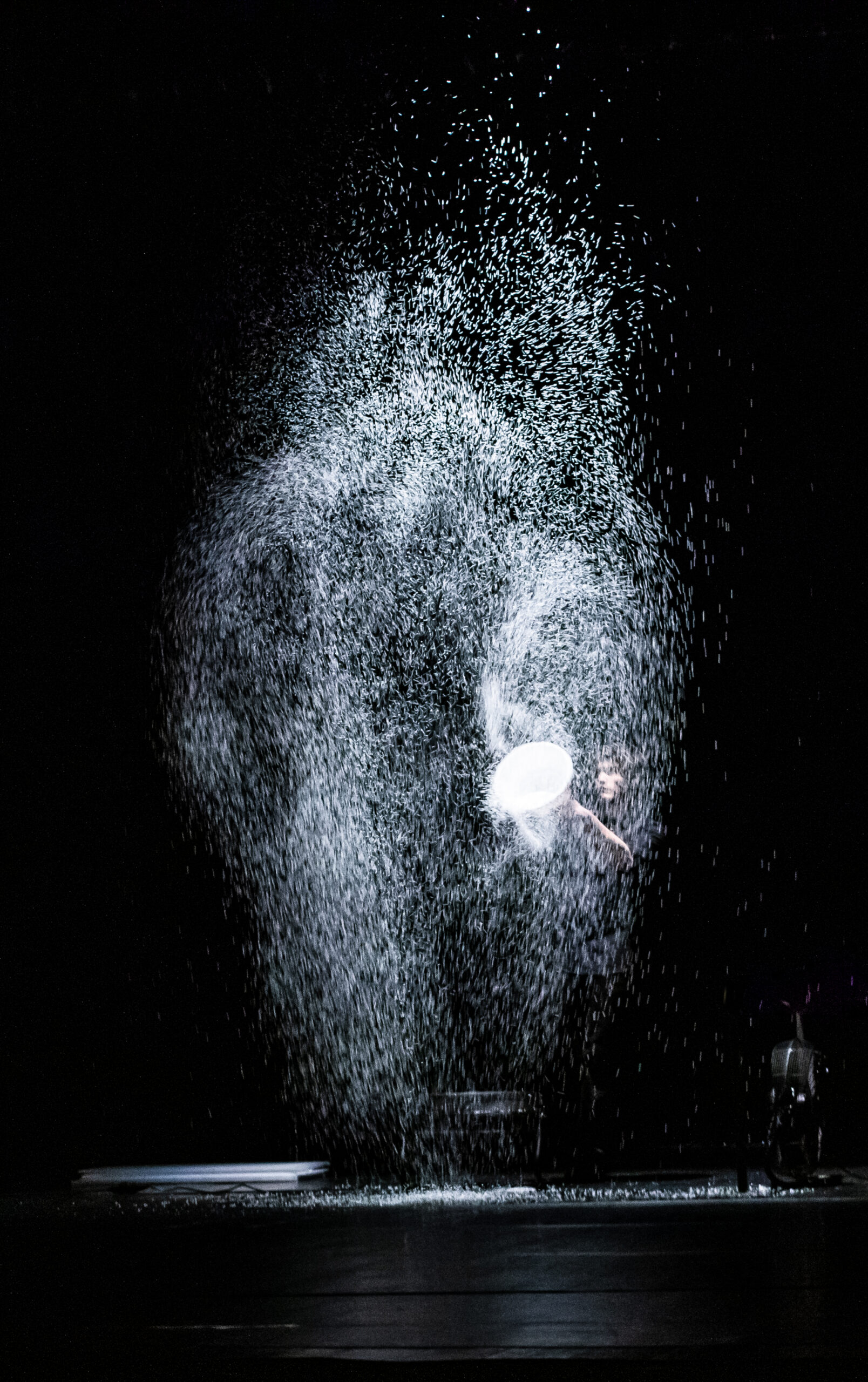 A photo of a person who has created a large cloud of white dust, perhaps polystyrene lit against a stage black out.