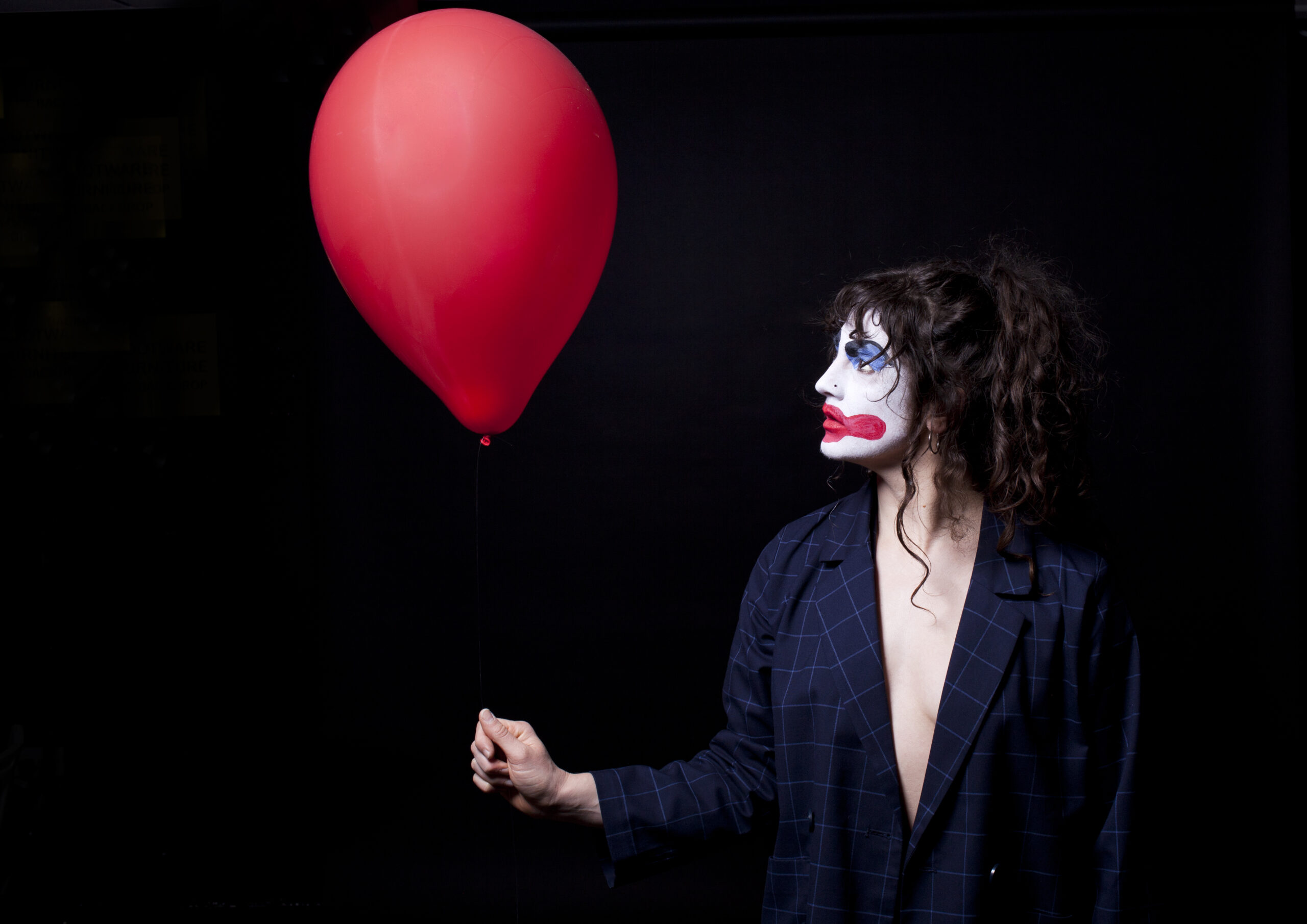 A photo of a person dressed in a suit jacket, holding a red balloon. They have traditional clown face make up on with their hair tied up. They are looking up at the balloon.