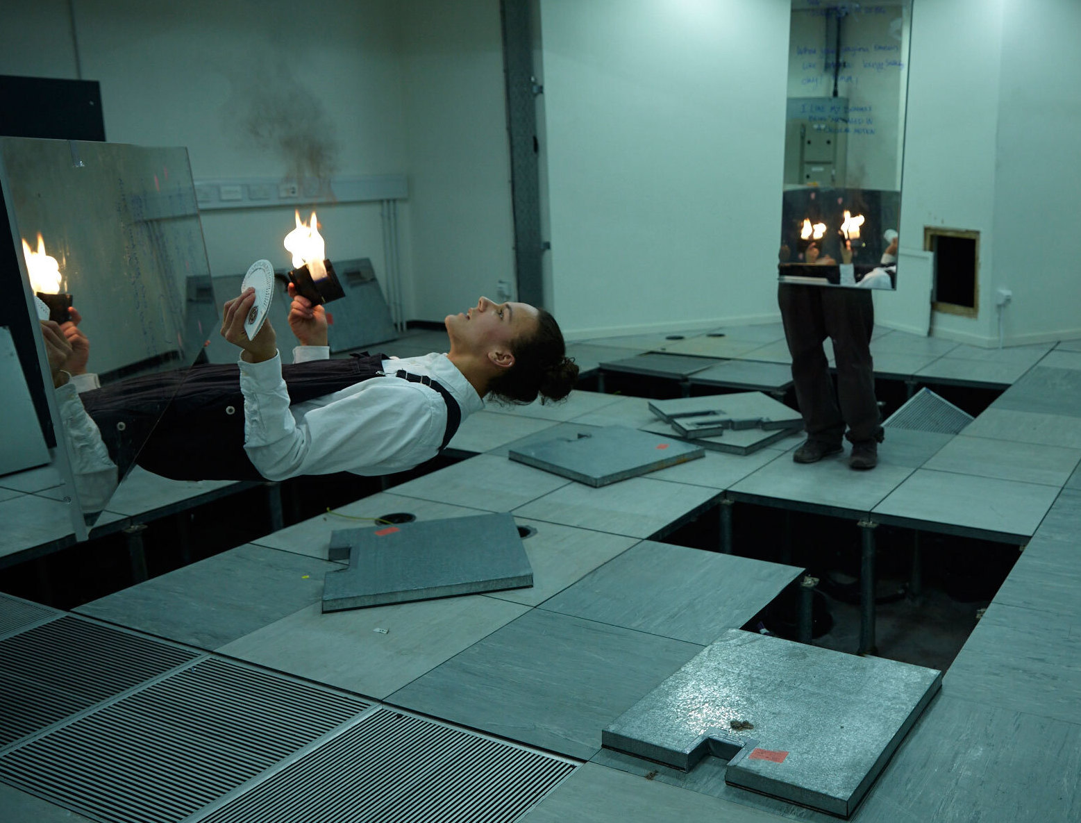 A person wearing a white shirt and black dungarees appears to be in two halves in an abandoned office room. The torso is horizontally disappearing into a mirror in the fore front of the image. The legs are standing vertically in the background with a mirror as the body, the mirror is reflecting the torso. The torso is holding an object on fire and a pack of cards spread in a fan.