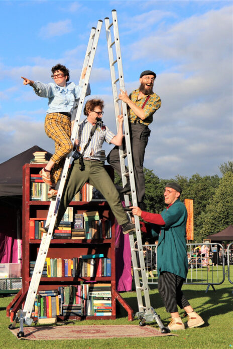 An image of Four performers up two ladders that are leaning together to make a tower. There is a performer balancing in between them and two performers are holding on to the outside whilst another performer holds the structure up. They are all looking in different directions. There is a large and colourful bookcase in the background.