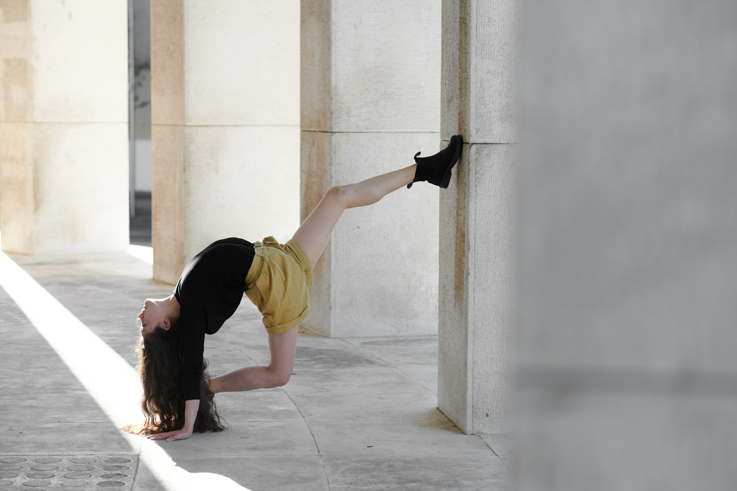 A photo of a woman outside in a paved area. She is doing a version of the splits but one of her legs is about head height on a pillar and her other on the floor, her torso is arching backwards and her head is nearly touching the floor behind her.