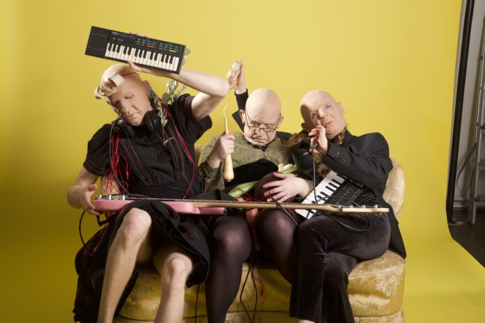 A photo of three people sat down on a yellow cusion. They rae wearing full face masks bold white men with glasses. They are playing a variety of electri instruments with wires dangling everywhere. One of the people is holding a parsnip and a leek and hitting a guitar with it.