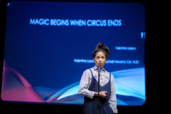 A photo of a person standing in front of a blue presentation screen talking into a microphone. The screen reads " Magic begins when Circus ends"