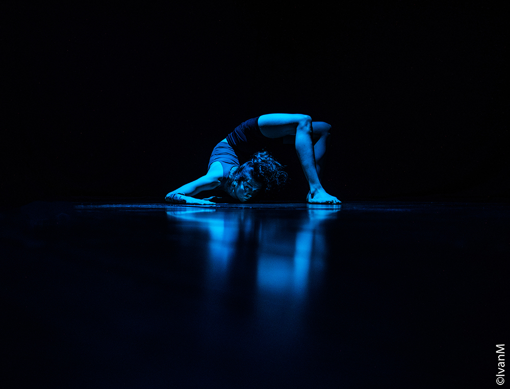 A photo of a person in a pool of blue light, they are in a contortion position where their head is nearly touching their heels on the floor.
