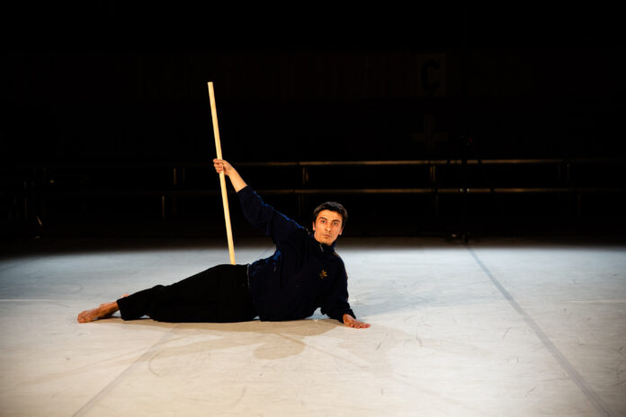 A photo of a man wearing all black, lying on a white stage. He is holding a stick about the length of his height, straight up in the air. He is looking directly at the camera.
