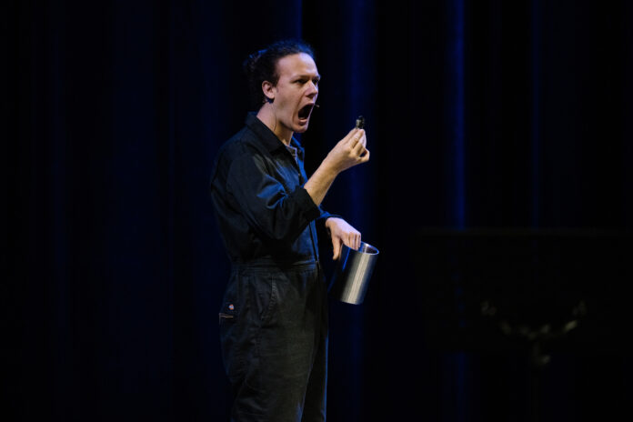 A photo of a person standing on stage in a black boiler suit holding a silver container in one hand holding a gold coin shaped object in the other. Their mouth is open as if they might eat the coin.