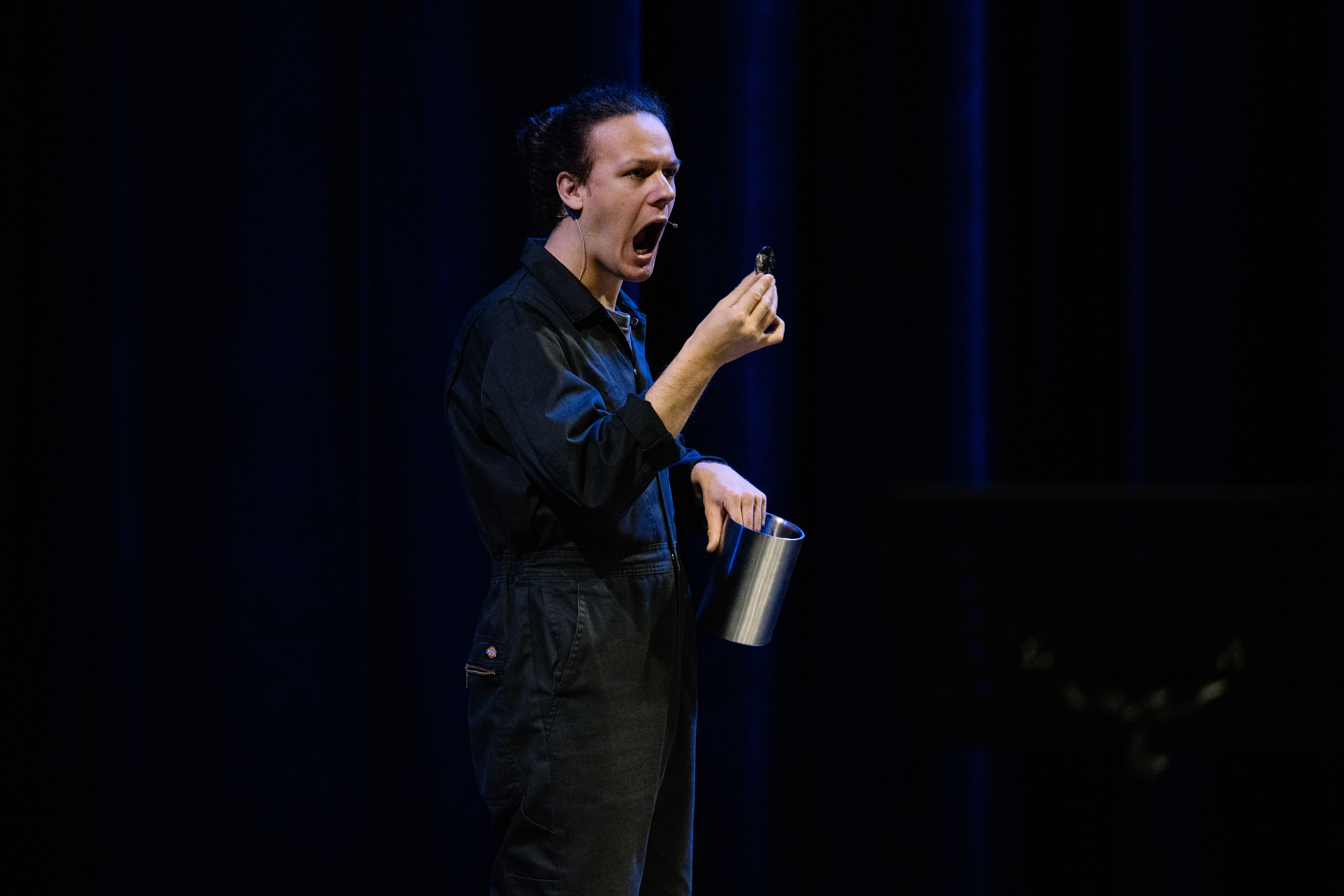 A photo of a person standing on stage in a black boiler suit holding a silver container in one hand holding a gold coin shaped object in the other. Their mouth is open as if they might eat the coin.